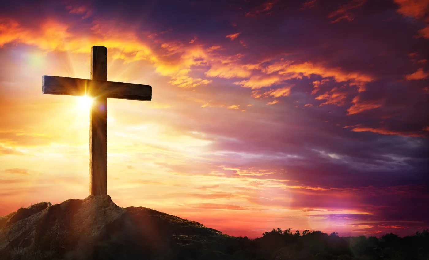 A wooden cross on a hill beneath a glowing orange and purple sky