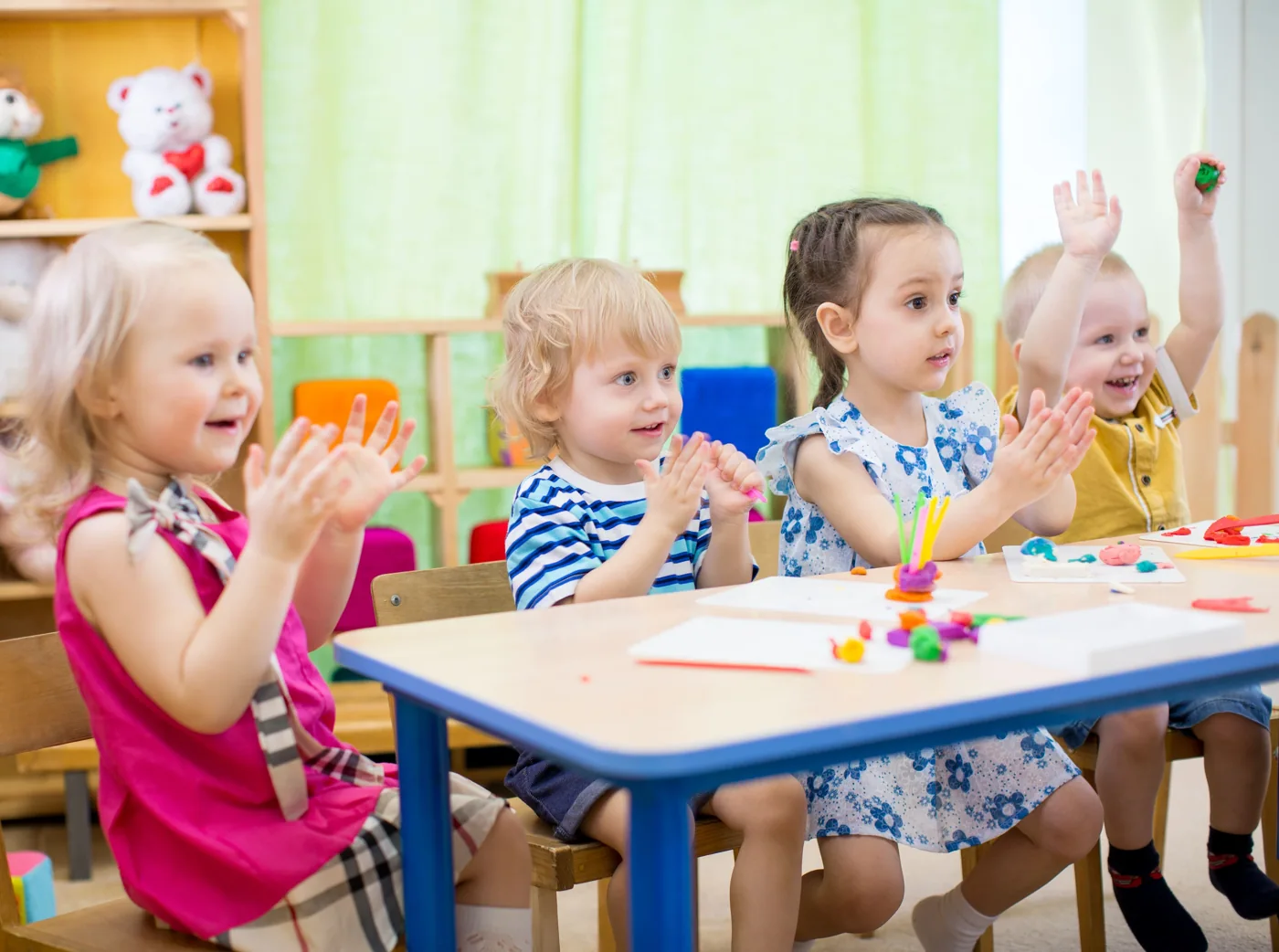 Young children clapping and exploring colorful modeling materials at a classroom table