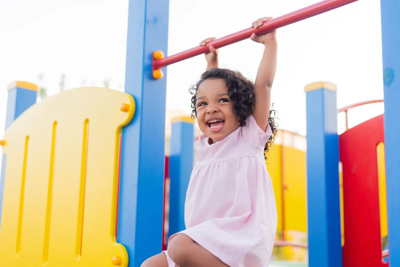 A smiling young child holding a red bar on colorful playground equipment