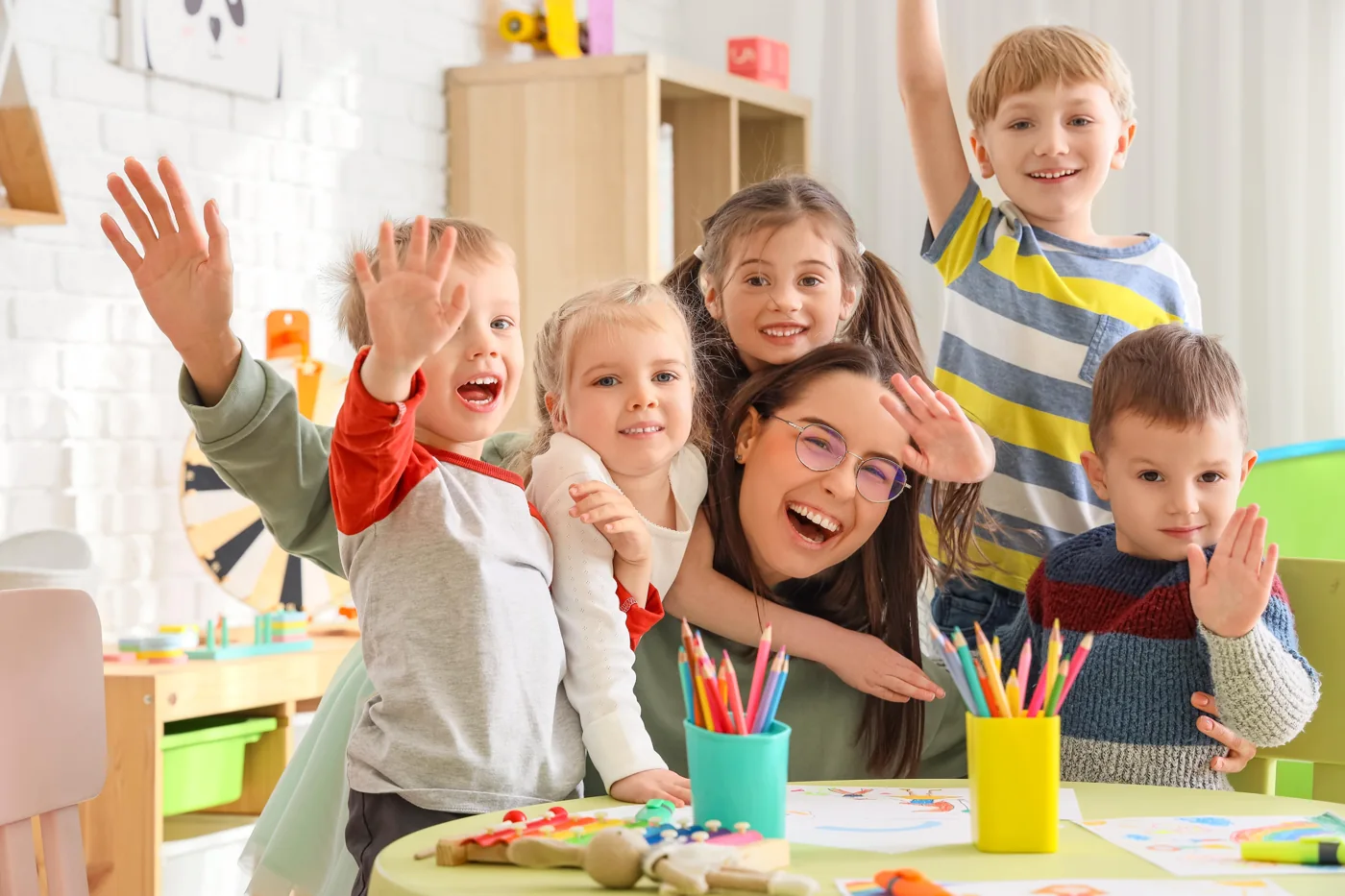 A smiling teacher and a group of children waving together in a classroom